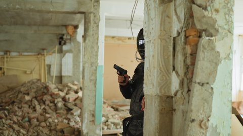 Fighter in black helmet with handgun looks from behind broken brick wall hiding from bad guys in old abandoned building corridor slow motion