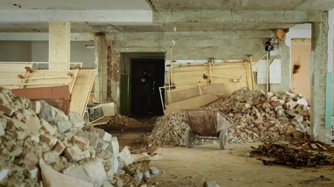 Army man in black moto costume with hardhat holds handgun coming into spacious hall with building rubbish in old abandoned premise slow motion