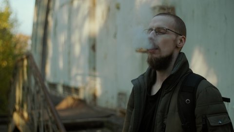 A bearded man in glasses smoking electronic cigarette blowing smoke and enjoying it. A vaping person with backpack standing by the porch outside looks like getting high. Blurred house background