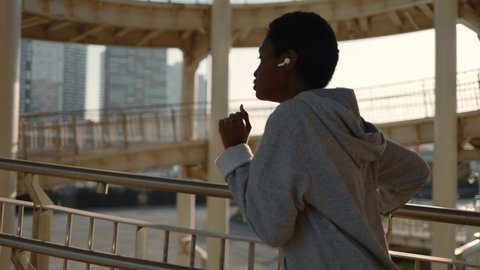 Healthy and fit african woman in wireless earphones jogging outdoors. Active young female dressed in sport clothes spending morning time for run on city bridge.