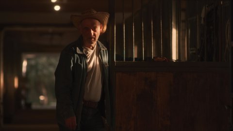 Aged man in straw hat standing in sunbeam near opened door of stall and looking at camera while working in barn on ranch.