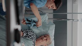 Vertical video: Medical assistant pointing at x ray scan for senior woman with illness in hospital ward bed. Nurse showing radiography to sick patient for healthcare diagnosis and exam results.
