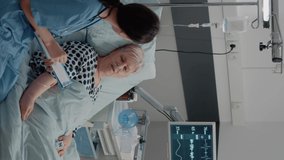 Vertical video: Nurse giving assistance to senior patient with disease in bed. Medical assistant and doctor doing healthcare checkup for pensioner with oxygen tube and IV drip bag in hospital ward.