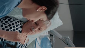 Vertical video: Woman nurse holding x ray scan to old patient for diagnosis and results. Medical assistant showing radiography to elder woman with disease for healthcare laying in hospital ward bed.