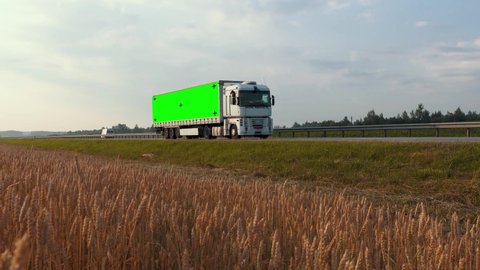 Truck with green screen and tracking markers on trailer drives along the highway past a wheat field in the rays of the dawn sun. Mockup concept for adding your logo, slogan or company name