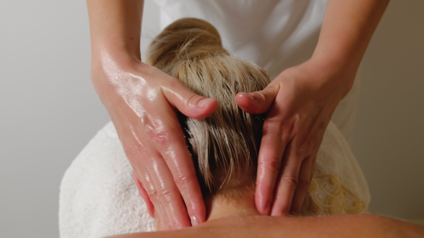 A girl massage therapist makes head massage to a young woman. Massage back of head and back of neck in salon. Therapeutic massage concept. Camera movement along body along back