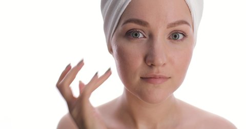 Portrait of a charming european young woman with a hair drying towel on her head applies skincare cream on face from her hand tapping and smiling for the camera on white background. Skin care concept.