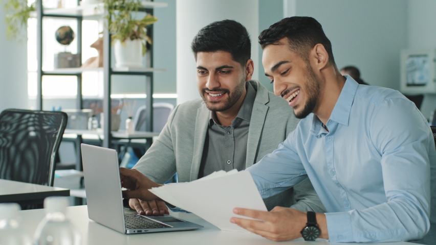 Cheerful positive friendly businessmen partners colleagues holding document contract looking at laptop screen discuss ideas having fun during break in office. Good business relationships, concept Royalty-Free Stock Footage #1081035323