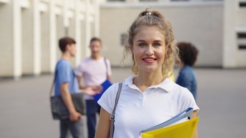 Blonde woman with curly hair and blue eyes wearing white polo shirt smiling at camera near college building on sunny day, group of people on blurred background. Tracking shot portrait of student