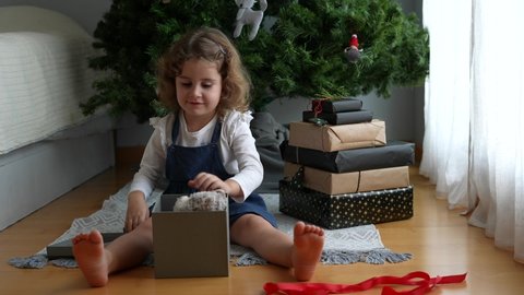 Little girl sitting next to opens the Christmas present and hugs the toy with illusion. Child girl with Christmas gift at home. Christmas surprise. Christmas holidays concept, New Years Eve