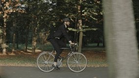 Businessman riding a bike in city park wearing helmet and a bag, side view. Businessman riding a bicycle in a public softwood park at the sunset. High quality 4k footage - Powered by Shutterstock - Get 15% off with code: PIKWIZARD15