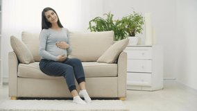 4k slowmotion video of pregnant woman sitting on white sofa and touching her stomach.