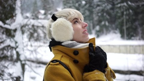 Girl in a yellow coat with earmuffs watching snow falling in the forest. Close up. 