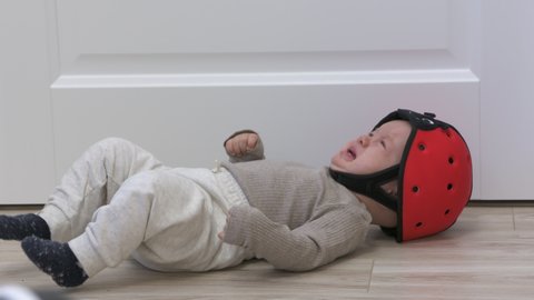 Baby boy wearing soft safety helmet falls backwards and hits his head on the floor, child crying lying on his back, head protector helps reduce impact of fall. High quality 4k footage
