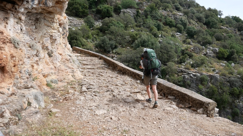 A girl tourist with a large backpack walks along the trail on a bright sunny day. Cliffs on the left, cliff on the right. The trail consists of ancient ruined steps.