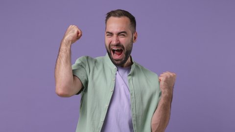 Excited jubilant overjoyed happy fun young bearded man 20s years old wears mint shirt doing winner gesture celebrate clenching fists say yes isolated on plain light purple background studio portrait