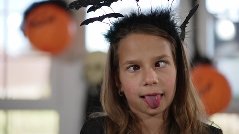 Headshot cute Caucasian girl in bat headband Halloween costume grimacing looking at camera crossing eyes. Smiling charming positive child making faces posing indoors on holiday. Slow motion