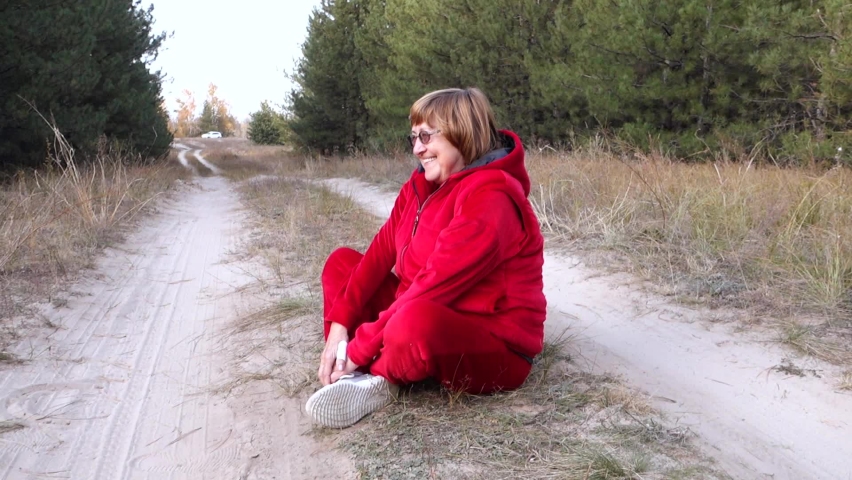 The red-haired grandmother in eyeglasses for sight laughs merrily, because she fell on a path in the forest and cannot get up.