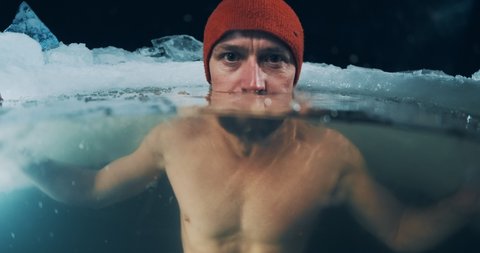Winter night swim and cold treatment. The young man soaks in the winter lake at night and looks at the camera. Split composition with an underwater view of the icy water