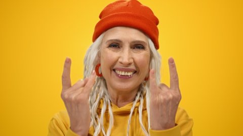 Smiling mature woman showing rock gesture on yellow studio background. Joyful retired old lady spending time inside. Cheerful elderly female person model face posing camera indoors. Happy retirement 
