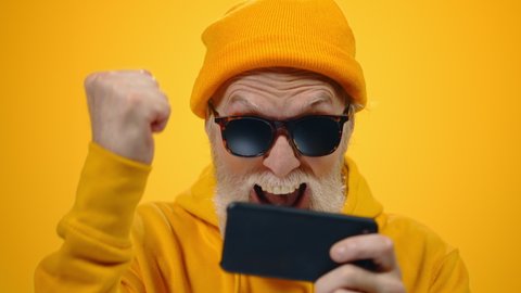 Aged happy man playing video game on cellphone inside. Excited old guy gesturing hands with smartphone on yellow studio background. Mature male person getting victory online on phone. Mobile app user
