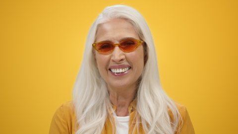 Happy senior woman in sunglasses posing in modern studio. Portrait cheerful elderly lady laughing on yellow background. Smiling old female person enjoying retired lifestyle. Happy retirement concept