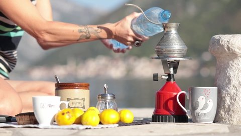 Female hands pour water from plastic bottle into coffee cezve making coffee outdoor on picnic at pier in bay of Donji Stoliv, Montenegro. Woman prepare coffee in Turkish cezve on camp gas burner