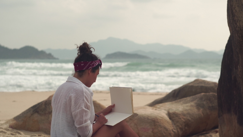 Young brunette woman sits on soft sand of wild beach writing with pen in blank notebook near rocks against raging sea in Vietnam closeup Royalty-Free Stock Footage #1081084064