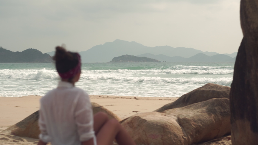 Young brunette woman in white shirt looks at raging sea under cloudy sky sitting near rocks on sand of wild beach in Vietnam Royalty-Free Stock Footage #1081084073