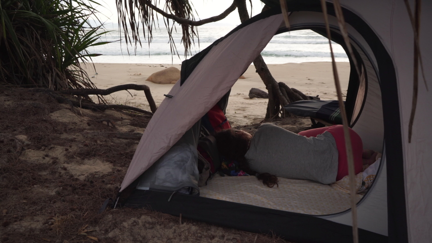 Brunette girl with braid lies in small camp tent on manless wild beach looking at waves in sea, under cloudy sky on gloomy day in Vietnam Royalty-Free Stock Footage #1081084091