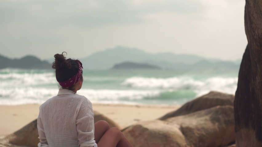 Young brunette woman in white shirt sits near rocks looking at raging sea with waves against mountains on wild beach in Vietnam Royalty-Free Stock Footage #1081084094