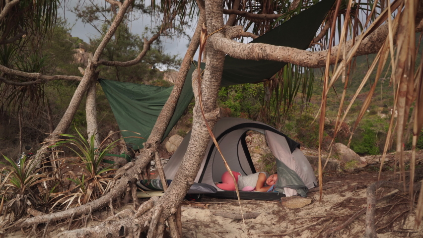 Young brunette girl with long hair sleeps calmly in small camp tent on manless wild beach under palm trees on gloomy day in Vietnam Royalty-Free Stock Footage #1081084097