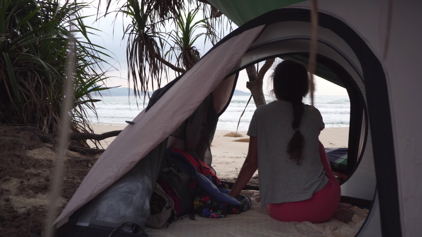 Brunette girl with braid sits in small camp tent on tropical wild beach looking at waves in sea under cloudy sky in Vietnam Royalty-Free Stock Footage #1081084100