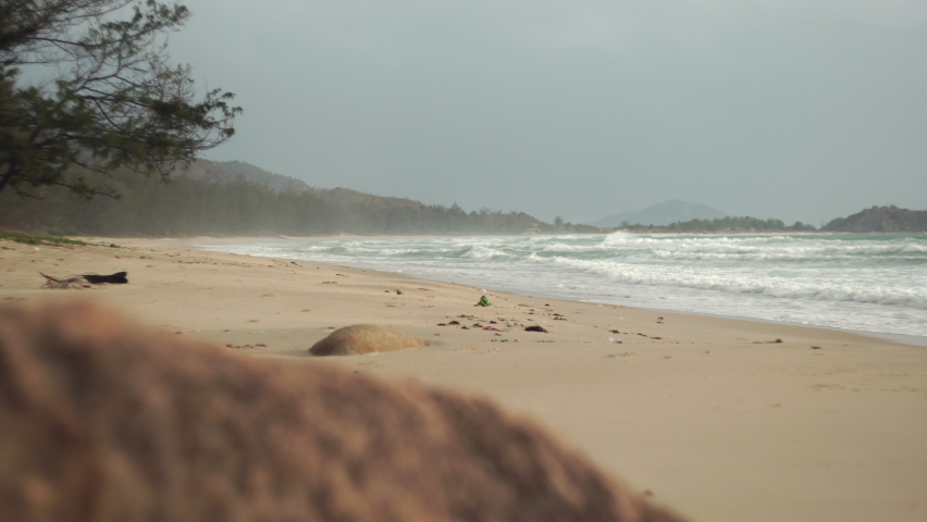 Low azure sea waves slowly wash over wild sandy shore under cloudy sky with rocks on foreground on quiet gloomy day in Vietnam Royalty-Free Stock Footage #1081084127