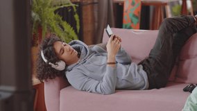 Teen boy lying on sofa, using smartphone and listening to music with wireless headphones while his sister sitting on floor and playing console game at home