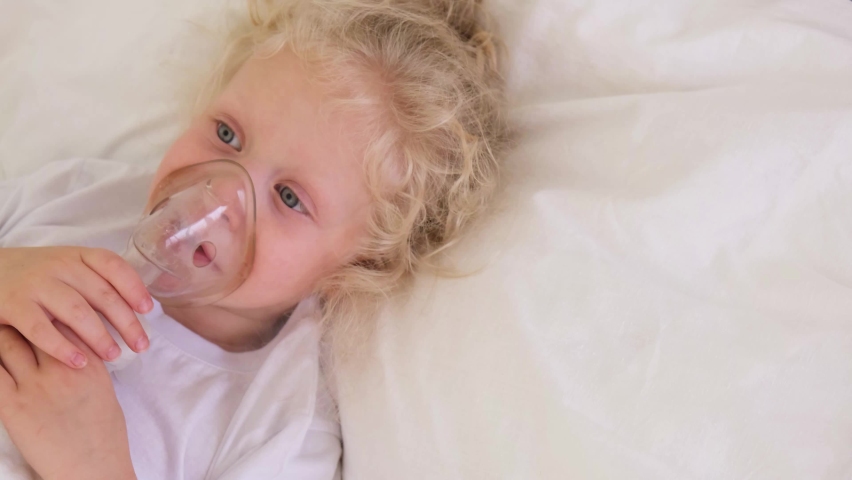Cute little girl with curly blond hair lies in bed, sick five-tape girl in bed makes inhalation with a nebulizer, treatment of bronchitis and lung disease in children | Shutterstock HD Video #1081086434
