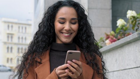 Happy latin hispanic girl young business woman walking in city street looking into mobile phone smartphone screen chatting online in net smiling answering message in social media using gadget device