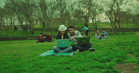 Beautiful young girl sitting on grass or lawn in the park and working on laptop . Boy coming asking to sit . Male and female students, friends talking , studying and discussing together . Slow motion