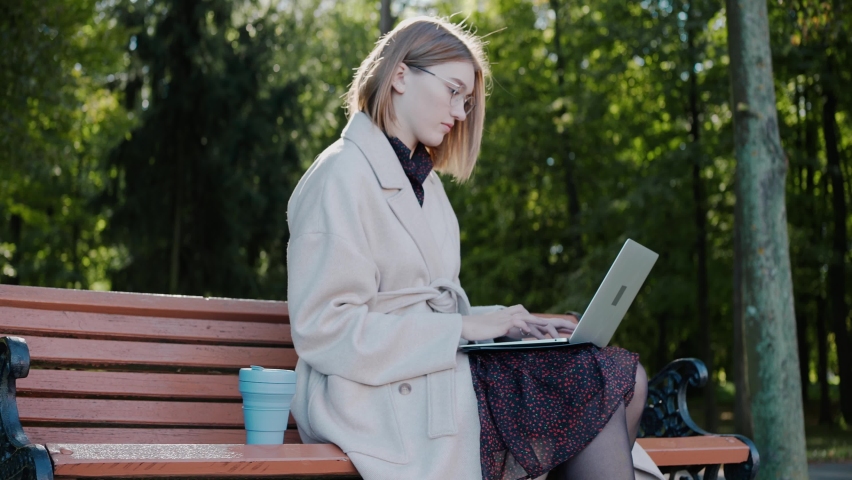 Young girl sitting on a park bench wearing a coat, writing on a laptop and drinking from an eco cup. Beautiful girl sits in the city park and works on a computer. High quality 4k footage Royalty-Free Stock Footage #1081097681