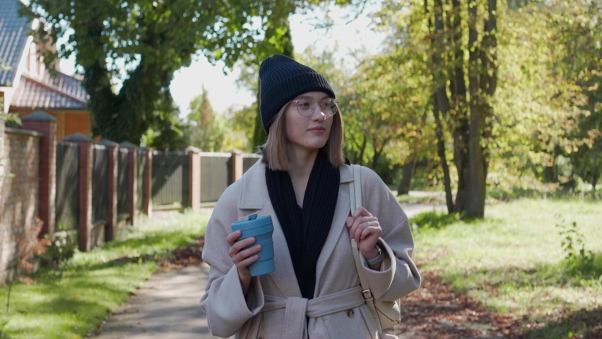 Young girl walks in the green neighborhood on a sunny autumn day, smiling and holding an eco cup. Beautiful girl walking in the park. Front view. High quality 4k footage
