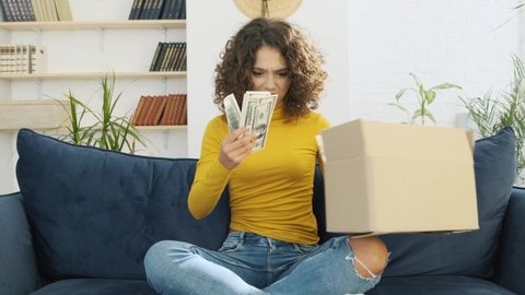 Disappointed young woman unpacking huge carton box full of dollar banknotes. Unsatisfied customer opening cardboard box with a cash