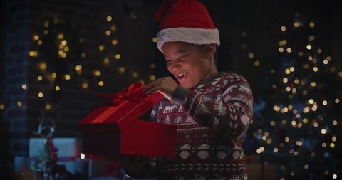 Little multiracial child boy in Santa hat opening gift box and wondering of the gift with Christmas Tree in the background