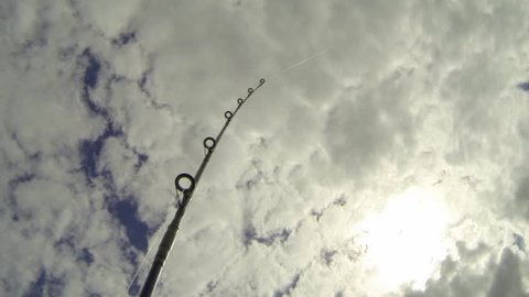Wide angle shot of a troll fishing rod bending and pointing towads sky with sunny clouds.
