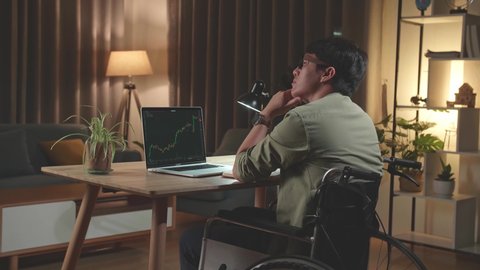Asian Man Trader Investor Sitting In A Wheelchair And Thinking About Something Then Raising Her Index Finger, Trading Data Index Chart Graph On Laptop Screen. Back View
