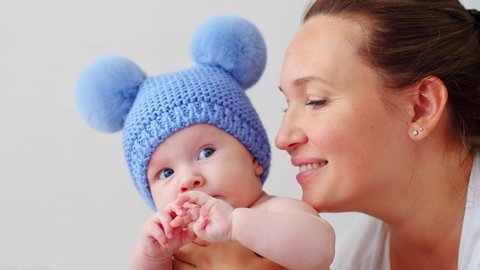 Happy Mother holds her Fun, Beautiful, cute little infant in a blue hat with pompoms .  Joyful Sweet newborn baby with his adult mother.  Two-month-old kid and adult mom. SLOW MOTION. Happy parent. 