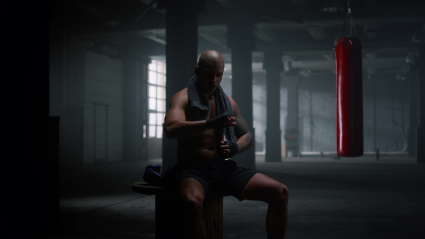 Muscular man professional boxer preparing for kickboxing fight. Powerful fighter wrapping hands boxing tapes in sport club. Multiethnic male kickboxer getting ready for strength exercises in dark gym Royalty-Free Stock Footage #1081140521