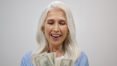 Happy senior woman counting banknotes on grey studio background. Rich old lady getting rich with jackpot inside. Successful mature female person holding cash money dollars. Wealth retirement concept