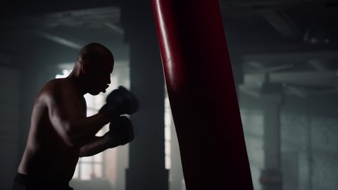 Professional boxer fighting punch bag in sports club. Shirtless man preparing fight. Multi ethnic fighter boxing training in dark gym. Close up strong body athlete silhouette. Power strength energy 