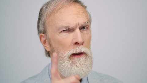 Mature serious man looking camera in studio grey background. Portrait focused old gentleman face wagging finger indoors. Close up disappointed male person frowning to camera inside.