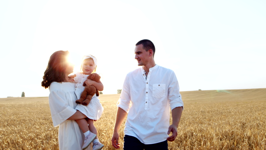 Cheerful couple walk across field: beautiful woman holds and carries little child, handsome man talks with kid. Spending leisure time together outdoors in nature, family summer stroll in countryside.